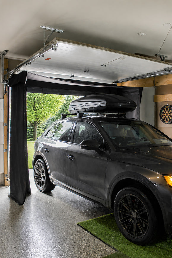 A dark gray SUV parked inside a garage with the door partially open, showing the car fitting under black curtains that are mounted on the ceiling.