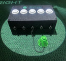 A black rectangular golf box with five white buttons on top, silver metal hooks on the front, and a green plastic cone attached to one hook by a black cord, placed on a green turf-like surface.