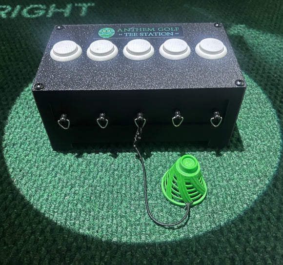 A black rectangular golf box with five white buttons on top, silver metal hooks on the front, and a green plastic cone attached to one hook by a black cord, placed on a green turf-like surface.