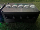 A black electronic control box with five white push buttons on top and five hanging loops on the front, placed on artificial green turf.