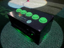 A black electronic golf tee system box with five green push buttons on top and green LED lighting on the bottom, placed on a textured green carpet.