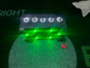 A rectangular electronic device with five clear illuminated dome hooks on top and multiple bright green LED lights glowing below, placed on artificial turf.