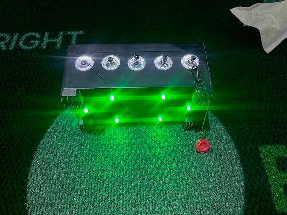 A rectangular electronic device with five clear illuminated dome hooks on top and multiple bright green LED lights glowing below, placed on artificial turf.