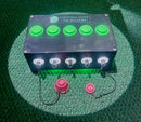 A black rectangular control box labeled 'ANTHEM GOLF TEE STATION' with five bright green buttons on top and five silver front connectors with lights, shown on a green turf-like surface with two small pink accessories below.