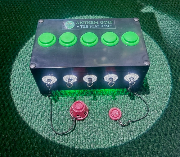 A black rectangular control box labeled 'ANTHEM GOLF TEE STATION' with five bright green buttons on top and five silver front connectors with lights, shown on a green turf-like surface with two small pink accessories below.