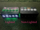 Two black electronic control boxes side by side on artificial grass, labeled 'Lighted' and 'Non-Lighted'. The left box has five green push buttons and glowing white lights underneath emitting green light. The right has five white buttons and no back lights.