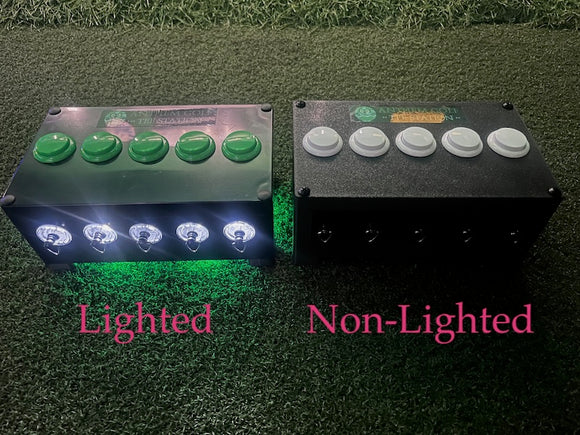 Two black electronic control boxes side by side on artificial grass, labeled 'Lighted' and 'Non-Lighted'. The left box has five green push buttons and glowing white lights underneath emitting green light. The right has five white buttons and no back lights.