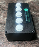A black control box with five white circular buttons on the front and a vertical label reading 'ANTHEM GOLF - TEE STATION' with a green logo, photographed on a speckled stone surface.