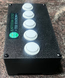 A black golf simulator target box with five white circular buttons on top and a green 'Anthem Golf - Tee Station' sticker on the side, placed on a speckled granite countertop.