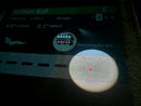 A close-up of a golf simulator screen showing a ball trajectory and a spotlight projection on the green, with the text 'Searching For Ball. On the floor is a black box with green buttons. attached to one of the front hooks is a golf tee