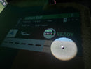 An indoor golf simulator screen and target mat showing a golf swing with a golf ball in flight, overlaid with course and score graphics including 'Anthem Golf' and a 'READY' status indicator. on the floor, a golf ball sits in a tee that is connected to a black box with five green buttons. 