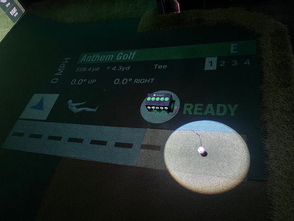 An indoor golf simulator screen and target mat showing a golf swing with a golf ball in flight, overlaid with course and score graphics including 'Anthem Golf' and a 'READY' status indicator. on the floor, a golf ball sits in a tee that is connected to a black box with five green buttons. 