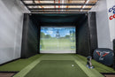 Indoor golf simulator setup with screen displaying a golf course and club equipment on a green floor.