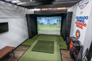 Indoor golf simulator setup with screen displaying a golf course, surrounded by golf bags and equipment.