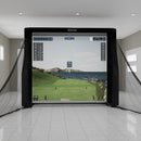 Indoor golf simulator with a large screen displaying a coastal golf course scene, surrounded by a net, in a spacious room with tiled flooring.