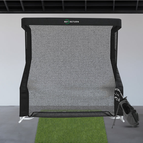 A golf practice net with a grass mat in front and a golf bag with clubs to the side, set in an indoor space.