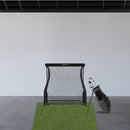 A golf hitting net set up indoors on a patch of artificial grass with a black golf bag and clubs next to it.