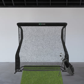 A golf practice net with a green mat and a bag of golf clubs stands indoors against a white wall.