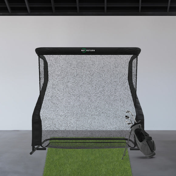 A golf practice net with a green mat and a bag of golf clubs stands indoors against a white wall.
